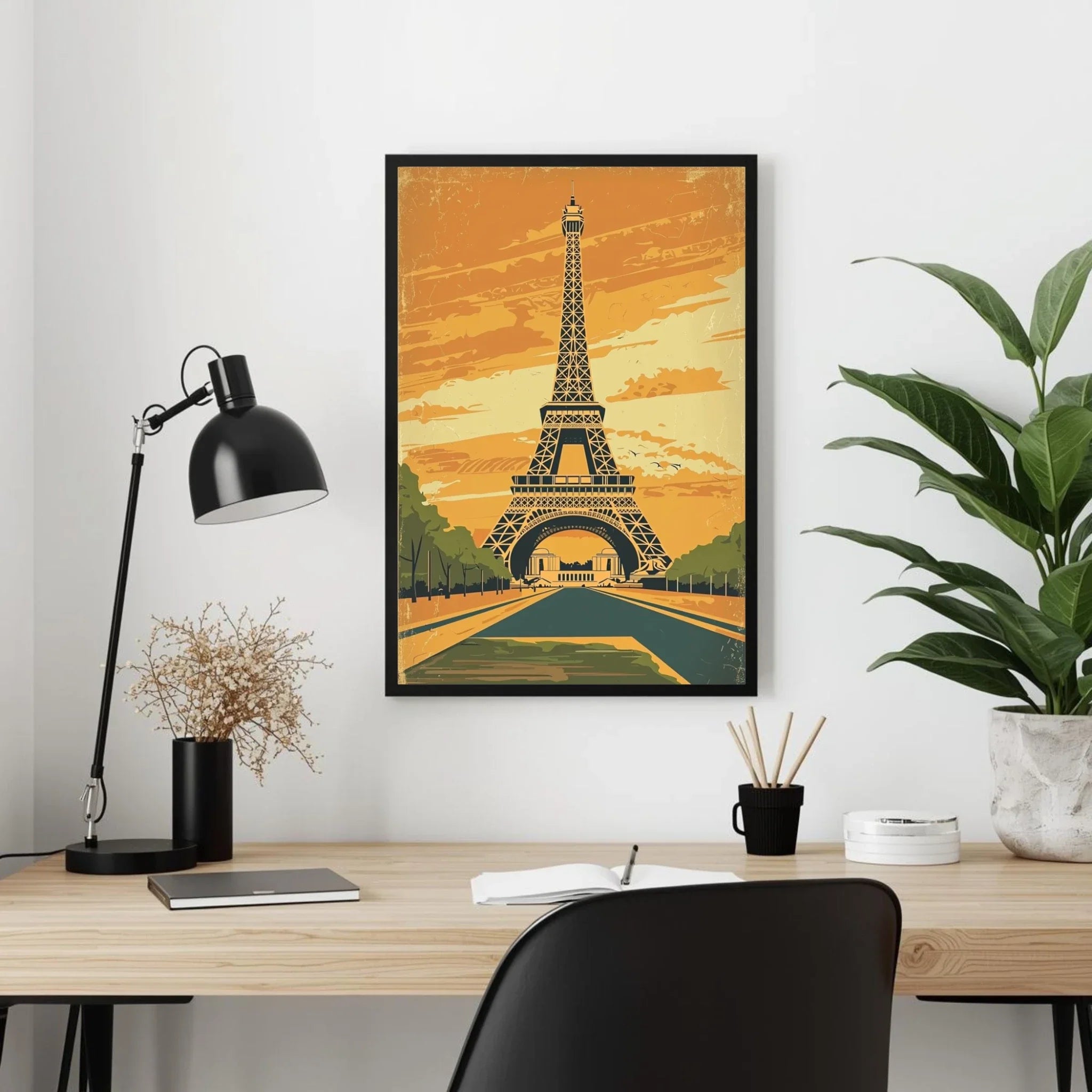 Framed Eiffel Tower poster on a wall above a desk with a lamp and plant.