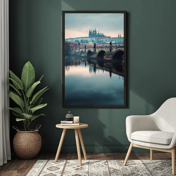 Framed artwork of a cityscape with a river and bridge, placed on a dark green wall above a small round wooden table with a plant and books.