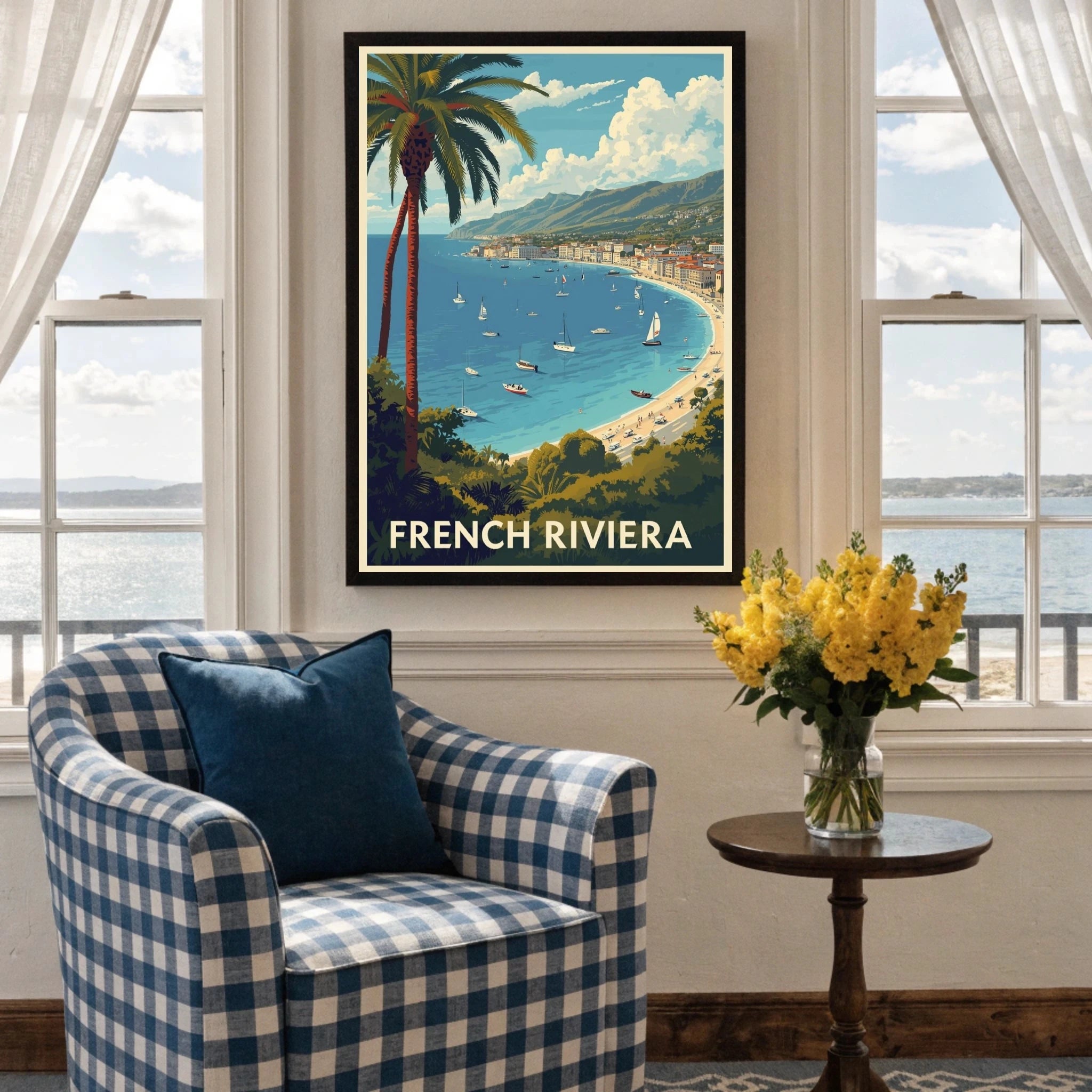 Framed artwork of the French Riviera above a blue checkered armchair with a table and flowers.