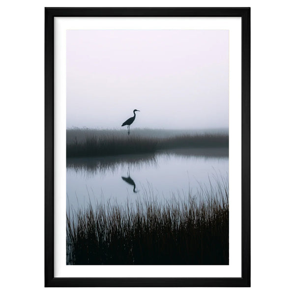 Solitary Grace minimalist nature artwork in a black frame leaning against an off-white wall, featuring a lone heron above mirrored water in soft early-morning fog.