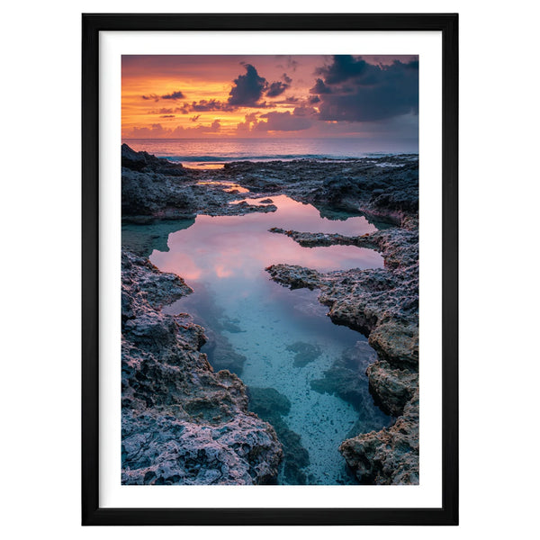 Tide of Light coastal artwork in a black frame leaning against an off-white wall, featuring soft sunset glow over rocky tide pools with coral and gold reflections on tranquil blue water.